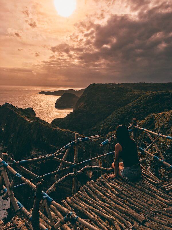 solo travel - girl sitting in tree house on top of mountain overlooking sunset in nusa penida